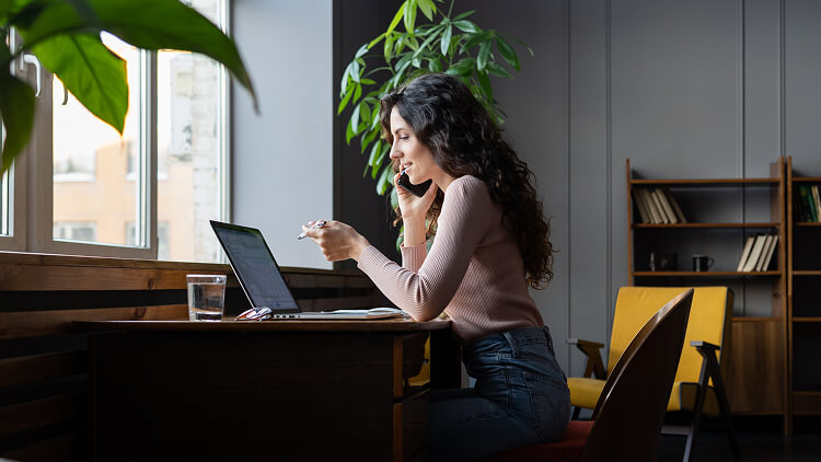 Persoon werkt flexibel op een laptop en telefoneert vanuit een moderne kantoorruimte met veel daglicht en planten.