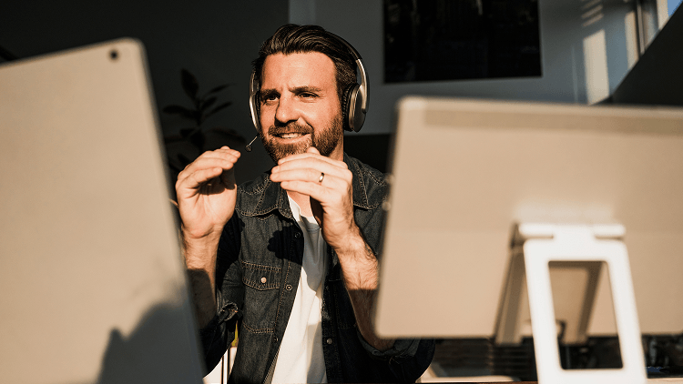 Man met headset zit achter twee computerschermen en voert een online gesprek in een kantooromgeving met invallend zonlicht.