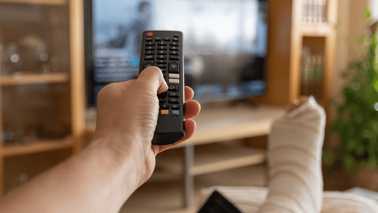 Close-up van een hand die een tv-afstandsbediening vasthoudt, met een verbonden voet op de voorgrond tijdens het televisiekijken thuis.
