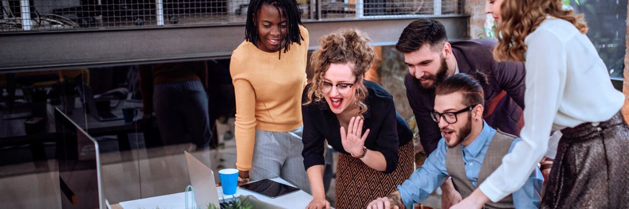 5 people at the office watching a laptop screen and the woman in the front is waving