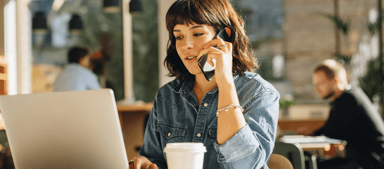 Vrouw belt met mobiel en zit achter laptop met koffie beker op voorgrond