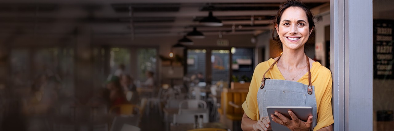 Woman in front of her business with a yellow t-shirt and a tablet in her hands.