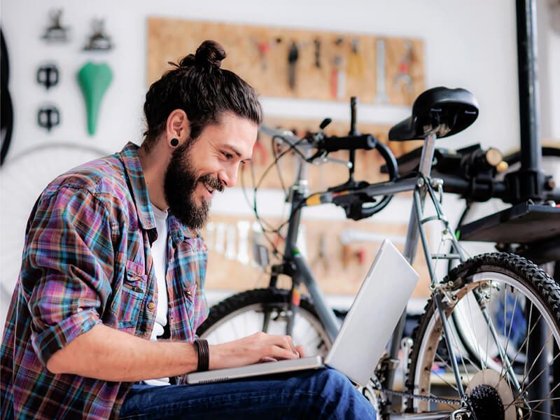 A man with a beard sitting down in a bike store with a laptop on his lap