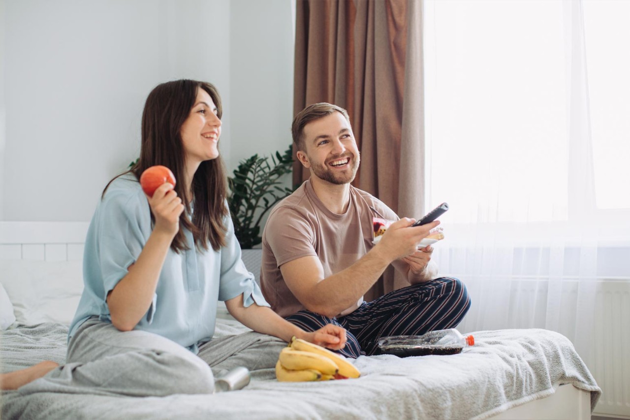 Happy young man and woman relaxing on bed at home watching TV together and eating donuts and fruit. The concept of rest and junk food.