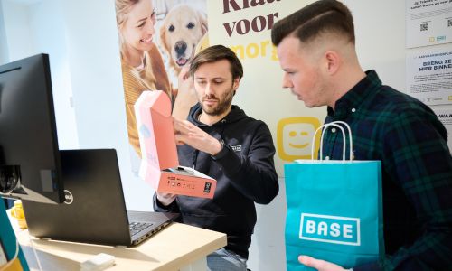 Two men in front of laptop and screen in Telenet shop with cellphone in box and Base shoppingbag
