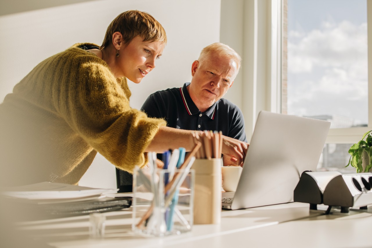 Manager pointing out laptop screen to colleague sitting at desk. Young female showing something on laptop display to mature man in office.