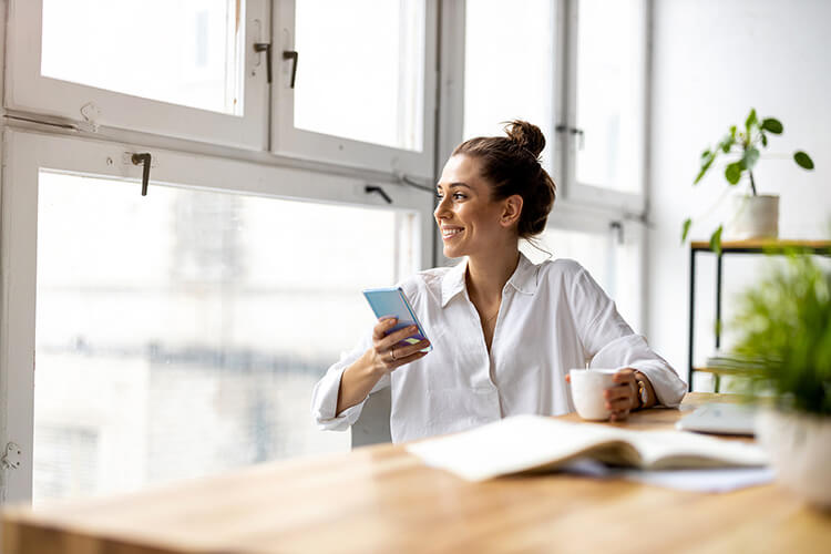 Woman sitting at the desk with a cup of coffee looking out the window with a smartphone in her hand